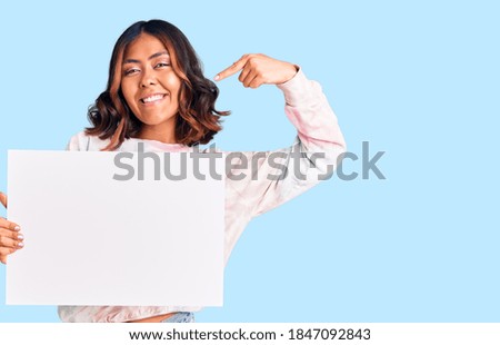 Young beautiful mixed race woman holding cardboard banner with blank space pointing finger to one self smiling happy and proud 