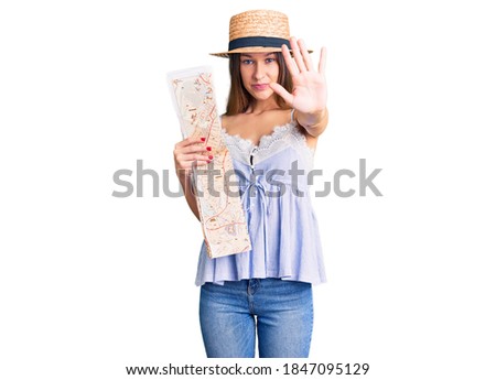Beautiful brunette young woman wearing summer hat holding city map with open hand doing stop sign with serious and confident expression, defense gesture 