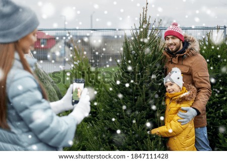 family, winter holidays and people concept - happy mother, father and little daughter buying christmas tree and taking picture with smartphone at street market over snow