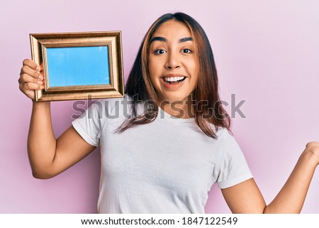 Young latin woman holding empty frame celebrating achievement with happy smile and winner expression with raised hand 