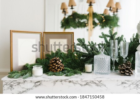 Christmas decor in home decoration: photo frame, spruce branches, glasses, candles and cones on a marble shelf