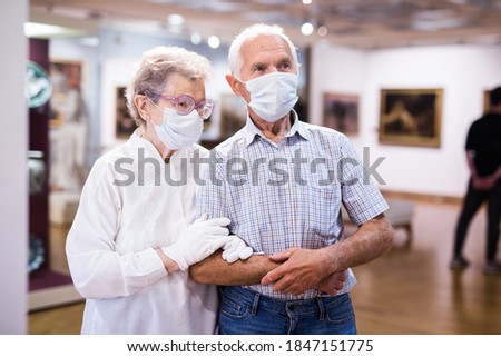 elderly European couple in mask protecting against covid examines paintings on display in hall of art museum