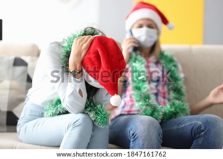 Woman in santa claus hat sits on couch with her head bowed next to woman in medical mask talking on phone. Pre-new year depression advice psychologist concept