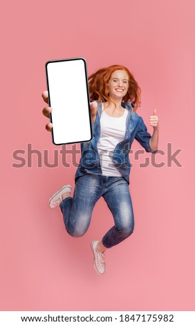 Cheerful girl redhead teenager jumping up, showing thumb up and newest smartphone with empty screen, enjoying new application for mobile phone. Pink studio background, creative image Royalty-Free Stock Photo #1847175982