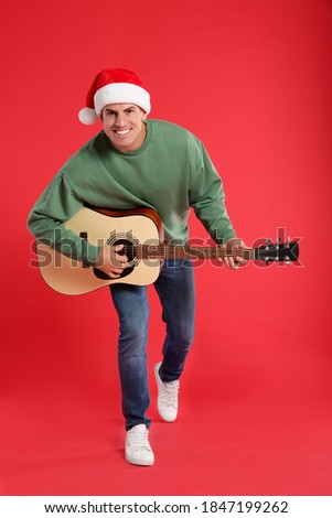 Man in Santa hat playing acoustic guitar on red background. Christmas music