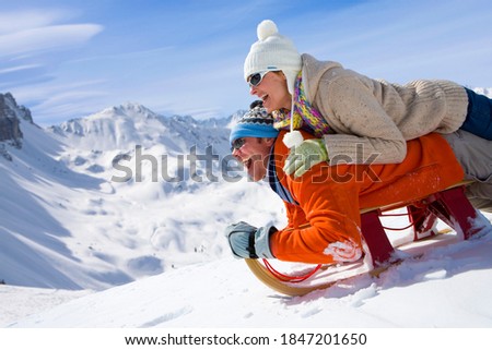 A side view of a happy couple having fun while sliding downhill together over snowy slope using a sled