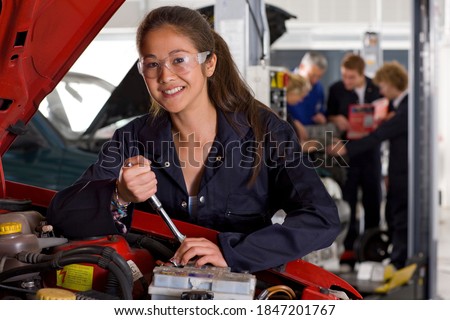 A horizontal view of a young girl in mechanic uniform and safety glasses repairing a car in the automotive training school while smiling at the camera Royalty-Free Stock Photo #1847201767