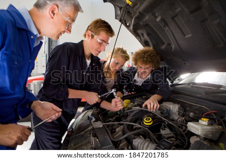 A side view of a young auto mechanics student repairing a car while being observed by his trainer and other students in an automotive training school Royalty-Free Stock Photo #1847201785
