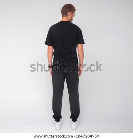 young stylish man in black t-shirt and black pants is standing from the back and looking away on the white wall background. medical concept. free space , mockup