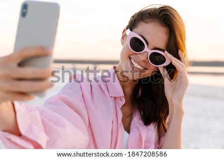 Cheerful young woman taking a selfie while standing at the beach during sunset, wearing sunglasses