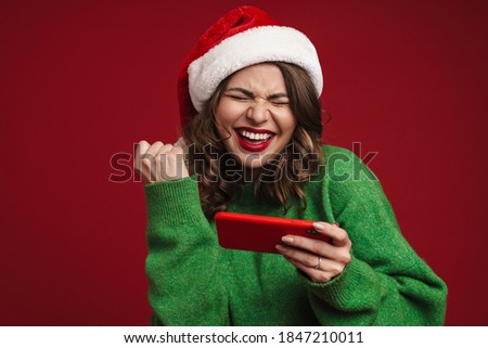 Photo of happy young woman in christmas santa hat using mobile phone and showing winner gesture isolated over red wall background