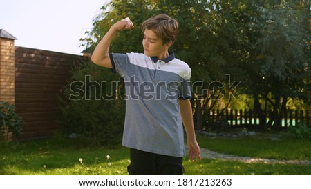A young handsome Caucasian boy stands on the street, shows a muscle on his right arm, knocks on it. Healthy lifestyle. Positive emotions. Royalty-Free Stock Photo #1847213263