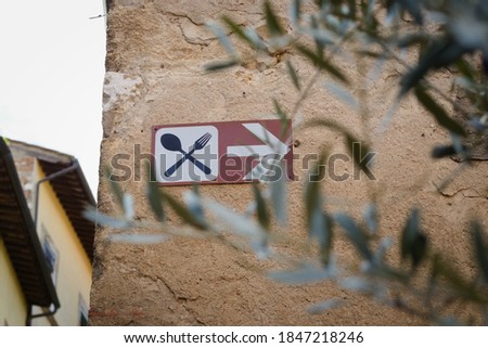 In focus, there is a sign on the wall with a fork and spoon and a pointer to the right - might be a restaurant. Out of focus olive branches in the foreground. It hangs on a beautiful warm colored wall
