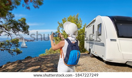 Caravan or towed trailer on seaside background. Female, girl, or woman tourist taking photo with phone or cellphone on vacation.