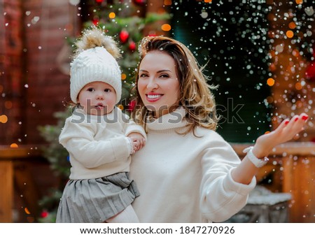 Young woman in white winter sweater posing with her toddler baby girl in the back yard with Christmas background. Snowing.