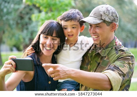 Happy excited military man, his wife and little son taking selfie on cell phone in city park. Front view. Family reunion or returning home concept