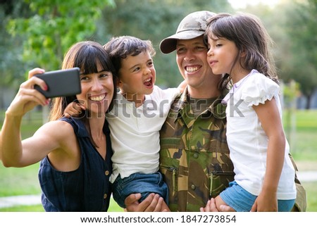 Happy joyful military family celebrating dads returning, enjoying leisure time in park, taking selfie on smartphone. Medium shot. Family reunion or returning home concept