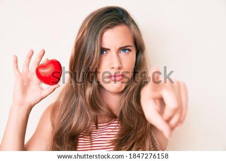 Beautiful young caucasian woman holding heart pointing with finger to the camera and to you, confident gesture looking serious 