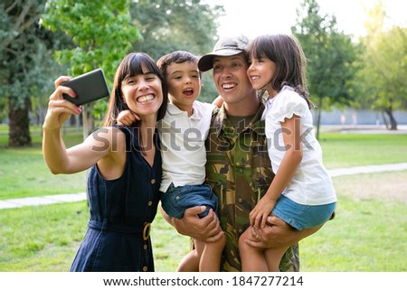 Happy excited military man, his wife and two kids celebrating dads returning, enjoying leisure time in park, taking selfie on cell phone. Medium shot. Family reunion or returning home concept