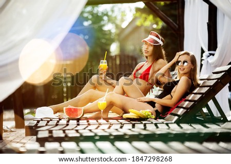 Two beautiful girls in swimsuits posing by wooden loungers with a dish of fruits