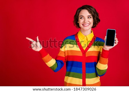 Photo of cheerful pretty lady direct finger look side empty space hold telephone offer low price sale smart phone device gadget wear stripes jumper cardigan isolated red color background