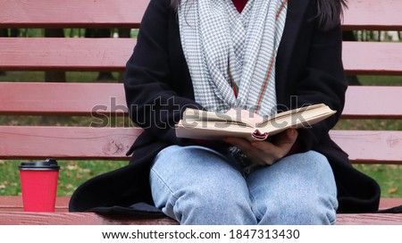 Young woman in jeans, coat and scarf, on a park bench. A woman is reading a book and drinking coffee or other hot drink outdoors alone. Close-up. The concept of honor, study, leisure and recreation.