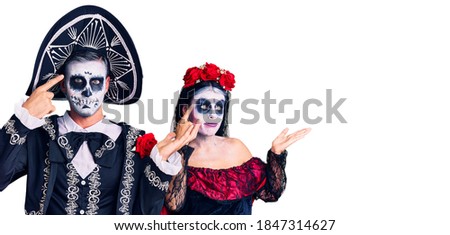 Young couple wearing mexican day of the dead costume over background confused and annoyed with open palm showing copy space and pointing finger to forehead. think about it. 