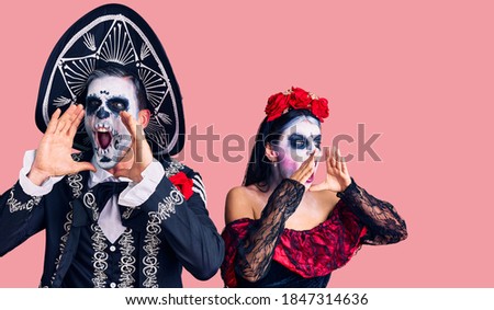 Young couple wearing mexican day of the dead costume over background shouting angry out loud with hands over mouth 
