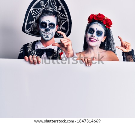 Young couple wearing mexican day of the dead costume holding blank empty banner smiling and confident gesturing with hand doing small size sign with fingers looking and the camera. measure concept. 