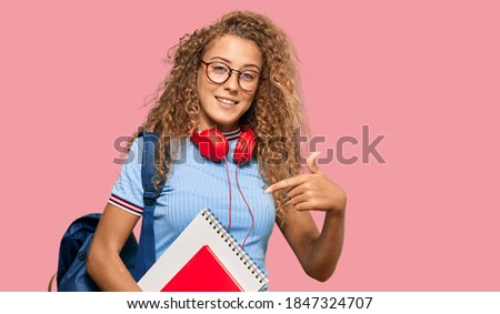 Beautiful caucasian teenager girl holding student backpack and books smiling happy pointing with hand and finger 