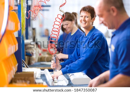 A medium shot of a young female worker smiling at camera while working on an aluminum light fittings on the production line with other workers. Royalty-Free Stock Photo #1847350087