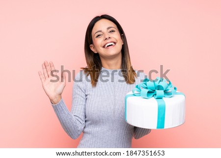 Woman holding a big cake over isolated pink background saluting with hand with happy expression