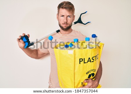 Young caucasian man holding recycling bag with plastic bottles and waste picker skeptic and nervous, frowning upset because of problem. negative person. 