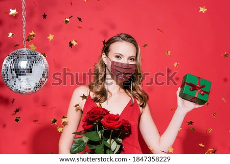 woman in protective mask with rhinestones holding christmas present near confetti and disco ball on red