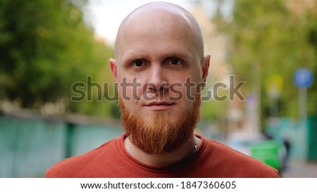 Close-up of the face of a young brutal guy with a bald head and a red beard with a serious face looking at the camera. Calm emotion. Summer evening. Royalty-Free Stock Photo #1847360605