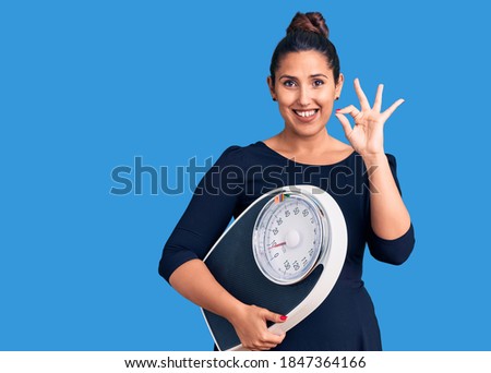Young beautiful woman holding weighing machine doing ok sign with fingers, smiling friendly gesturing excellent symbol 