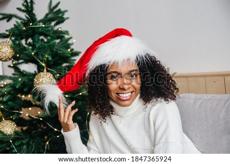 Portrait of smiling african american young woman in santa hat sitting at home onchristmas tree background. Celebrate Christmas and New Year at home.