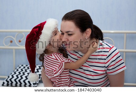 Little daughter in a hat of Santa Claus kisses her mother. Happy New Year.