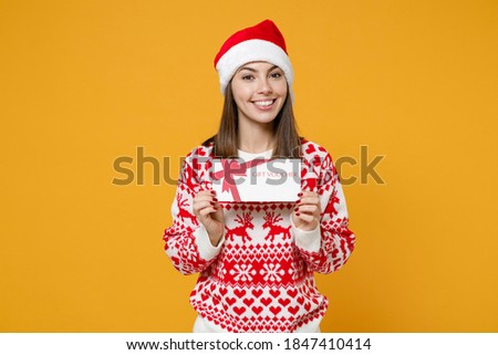Smiling young brunette Santa woman 20s wearing red sweater Christmas hat hold gift certificate isolated on yellow colour background, studio portrait. Happy New Year celebration merry holiday concept