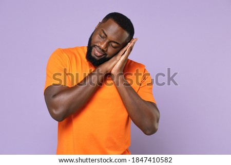Relaxed young african american man 20s wearing basic casual orange t-shirt standing sleep with folded hands under cheek keeping eyes closed isolated on pastel violet colour background studio portrait