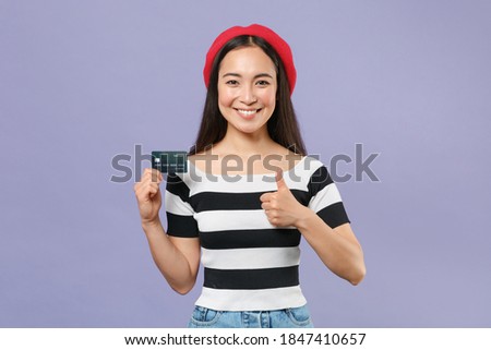 Smiling funny young brunette asian woman 20s wearing striped t-shirt red beret standing holding in hand credit bank card showing thumb up isolated on pastel violet colour background studio portrait