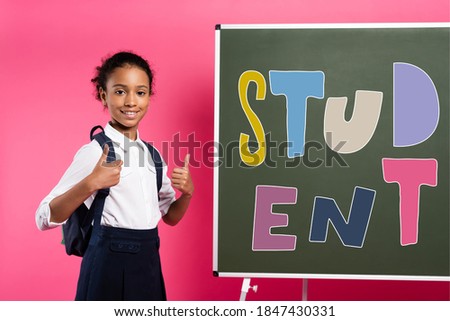 african american schoolgirl standing near chalkboard with student lettering and showing thumbs up on pink