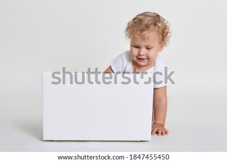 Cute little boy with white laptop computer, looking at screen with interest, child with curious facial expression sitting on floor, wearing t shirt, infant watches cartoons.