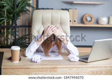 Tired young business woman feeling bad because of stressed workday. Holding head in hands and leaning on the desktop near crumpled paper and laptop in the office Royalty-Free Stock Photo #1847496070