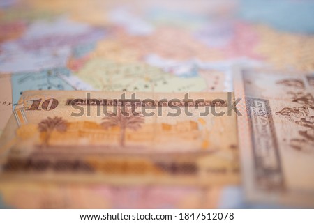 Close up picture of a Ten Saudi riyals bill next to a Kuwait Quarter Dinar, both below Saudi Arabia on a colorful and blurry map of Western Asia