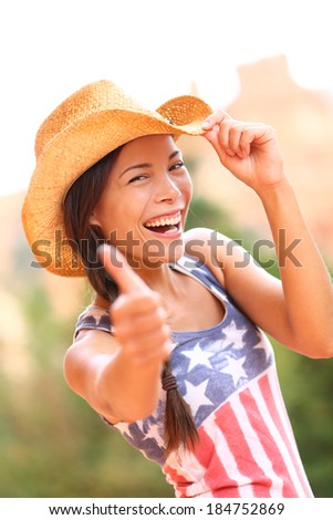 American cowgirl woman happy excited giving thumbs up wearing cowboy hat outdoors in countryside. Cheerful elated joyful woman smiling enjoying freedom. Beautiful multiracial Asian Caucasian female.