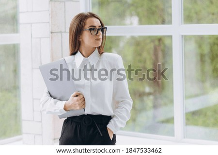 young successful girl standing in the office and holding a laptop. The girl is dressed in a white shirt and black pants