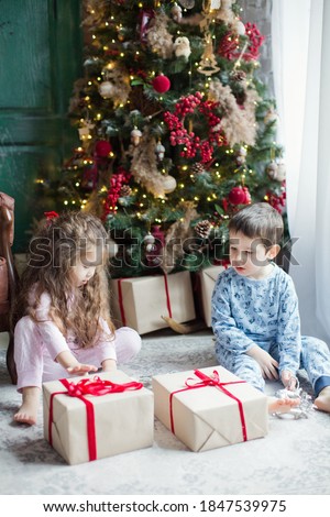 Toddler boy and girl in pyjamas hold gift boxes near Christmas tree in light Christmas interior. Cute Christmas kids