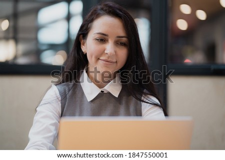 portrait of a Brunette Caucasian appearance in a white shirt office employee young woman smiling and sending emails. sitting in a chair and typing text on a laptop, in a coworking