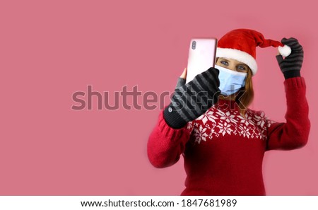 A young girl in santa claus hat and medical protective mask takes a selfie on her phone camera. Congratulations during self-isolation, coronavirus pandemic